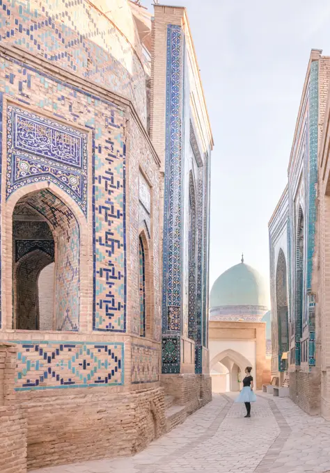 Female tourist walking on sidewalk near aged ornamental building of Shah-i-Zinda necropolis on sunny day in Samarkand, Uzbekistan