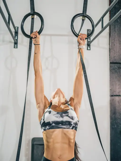 Crop strong adult female athlete in gray sportswear working out on black gymnastic rings against white wall in sport club