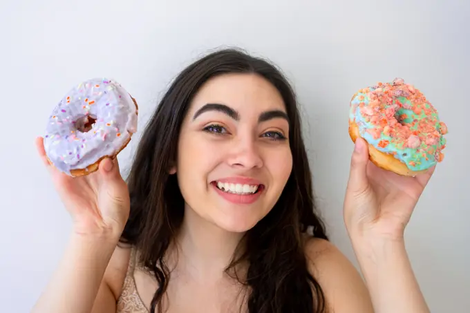 Carefree woman having fun and playing with glazed pastry with sprinkles while standing by white wall