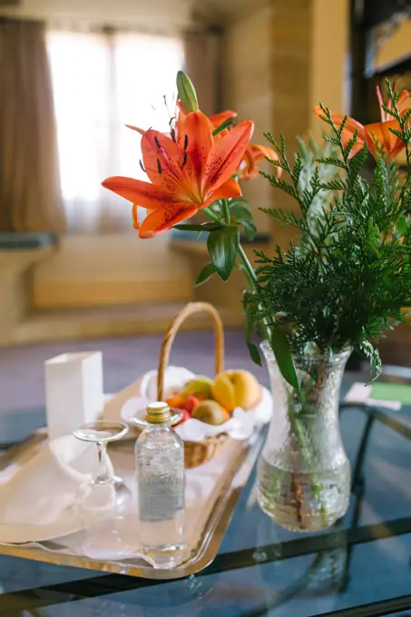 Vase with beautiful flowers and tray with breakfast food placed on glass table in luxury hotel room in morning