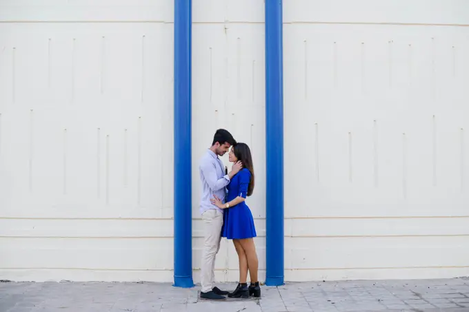 Side view of loving man and woman in elegant clothes kissing on background of white wall with bright blue columns