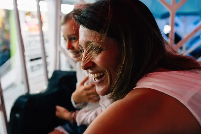 Cheerful adult woman smiling and looking away while riding Ferris wheel with daughter in amusement park