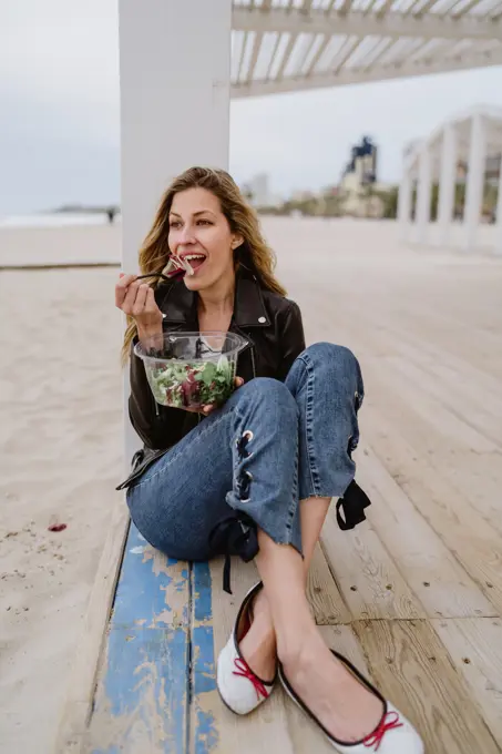 Stylish long haired blonde woman in black jacket enjoying healthy green salad while sitting on wooden terrace on coast smiling looking away