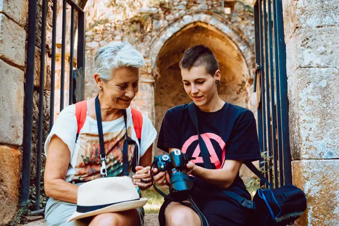 Enthusiastic grandmother and granddaughter sitting at stony entrance of ancient building with metal doors and watching photo pictures on camera
