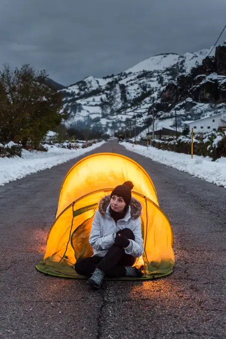 Woman in winter clothes sitting at entrance of tourist tent illuminated from inside standing in middle of asphalt road in mountain village at evening