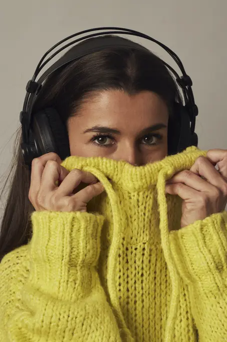 Cheerful young dark haired female in headphones looking at camera while covering face with yellow cozy sweater on gray background in studio;Playful young woman listening to music in studio