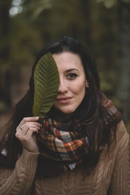 Stylish lady in knitted sweater and checkered scarf covering eye with leaf while standing on blurred background of forest;Woman covering eye with leaf