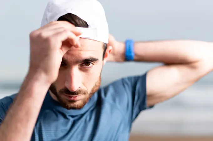 Sportsman in blue t shirt with hands behind head on white cap looking at camera with empty sandy seaside on blurred background