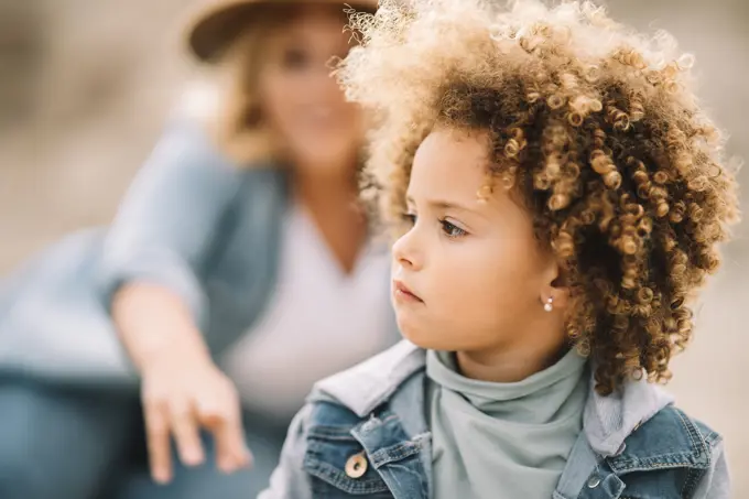 Concentrated serious toddler with curly hair dressed in casual clothes sitting on nature and looking away while woman resting behind and smiling;Focused curly child resting with mother