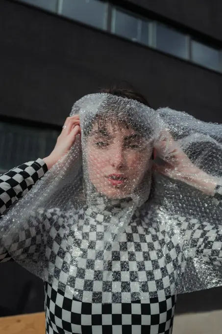 Emotionless female model in stylish black and white plaid jacket standing and wrapping face in bubble wrap while looking at camera at city street ;Fashionable woman in trendy casual wear in bubble wrap at city street