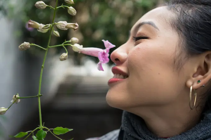 Closeup of crop Asian female on vacation smiling while enjoying smell of purple flower with lane on blurred background at Albaicin in Granada;Charming Asian female on vacation sniffing flower at city street