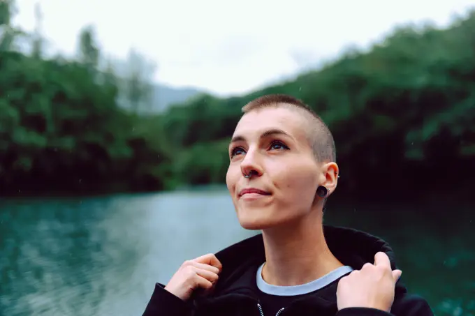 Young extraordinary woman with short hairstyle and piercing in casual clothing looking up to sky with pond among green plants on blurred background