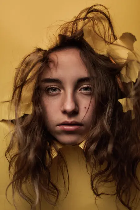 Pensive happy youthful female teen with uncombed hairstyle watching out from ripped hole in yellow paper in studio;Thoughtful female teenager looking out from hole in studio