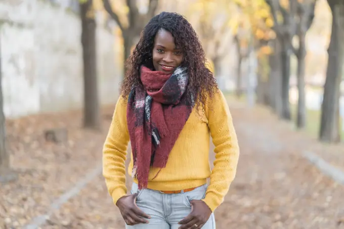 Charming fashion African American woman in a winter scarf with hands in pocket on road with autumn leaves in park