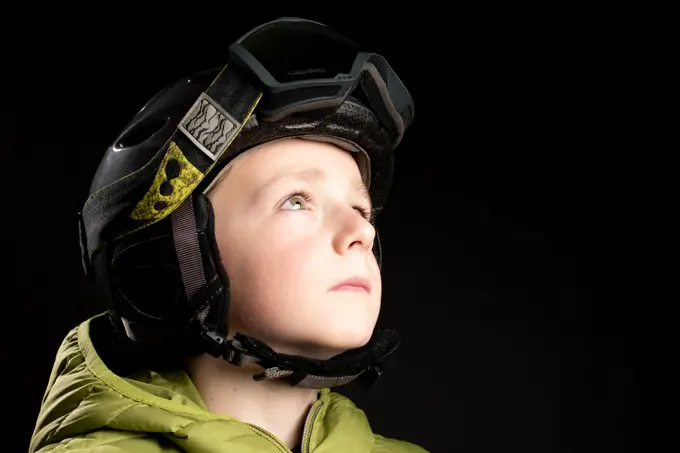 Focused little sportive kid in winter clothes with ski goggle on black background in studio looking away