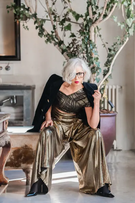 Calm senior confident lady in glasses and elegant clothes looking at camera while sitting on shabby bathtub edge among rich retro interior including old furniture and big green plotted tree in spacious room of country house