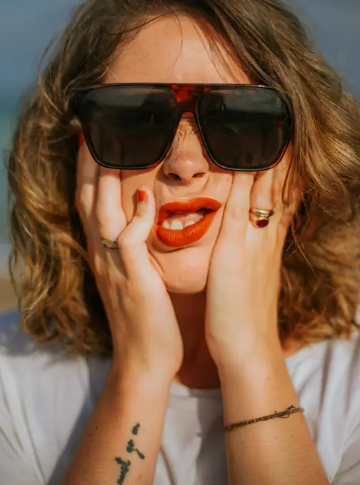 Stylish brown curly haired woman with red lipstick in trendy sunglasses looking at camera while squeezing face in palms;Contemporary resting lady touching face