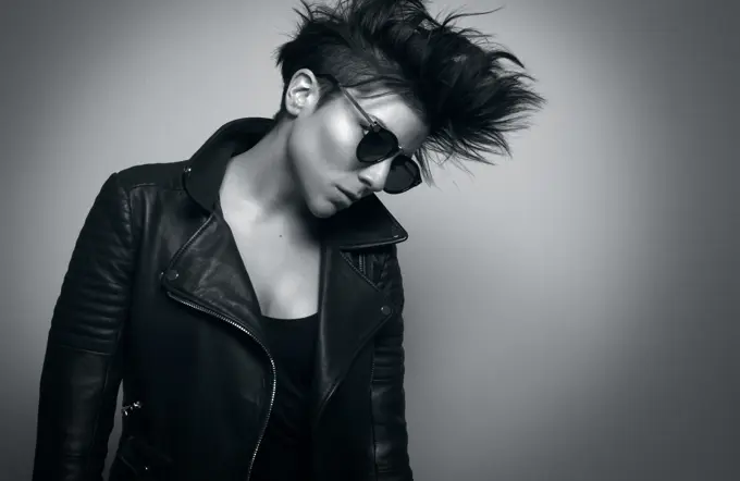 Independent serious youthful woman with short dark hair and sunglasses wearing biker jacket and looking down in studio on gray background;Strong young female rocker in leather jacket in studio