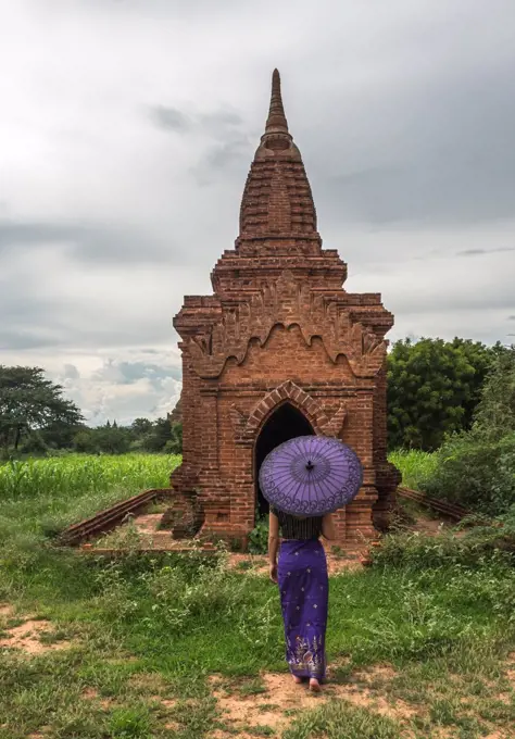 Back view of female in long dress holding traditional Burmese purple umbrella while standing on green meadow in front of old stone temple in ancient city Bagan in cloudy day;Woman with umbrella standing in front of ancient temple