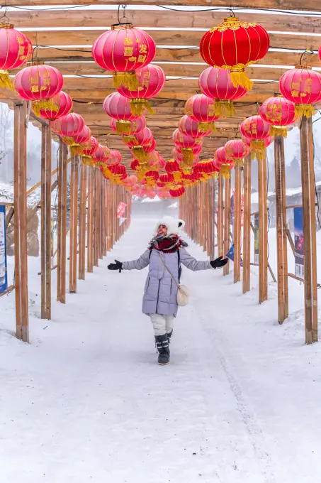 Full body happy young female with outstretched arms admiring red Chinese lanterns while walking in wooden passage on winter day;Cheerful woman walking in archway with Chinese lanterns