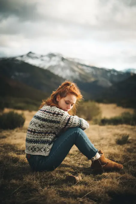 Side view of pensive charming woman with red hair in warm sweater sitting with closed eyes on lawn in mountain in gray day;Red haired woman thoughtfully sitting on meadow in mountains