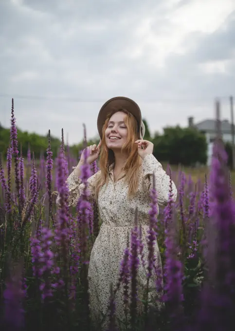 Happy sensual tender redhead lady in trendy hat with closed eyes enjoying smell of salvia blossom in meadow of Asturias, Spain;Romantic women in salvia field