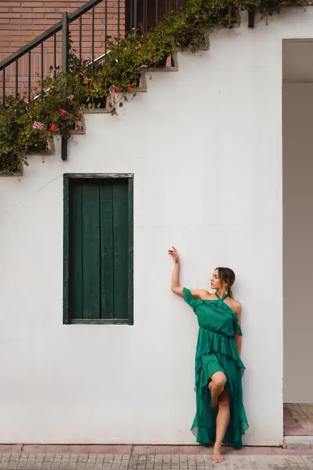 Full body barefoot female in green dress leaning on white wall of house with staircase and potted flowers on town street