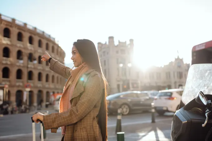 Side view of Asian female with suitcase smiling and stretching out arm while hailing cab on street of tourist city