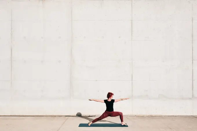 Side view of trendy fit female athlete in sportswear doing warrior two yoga pose on sports mat while training alone on street against concrete wall in sunny day