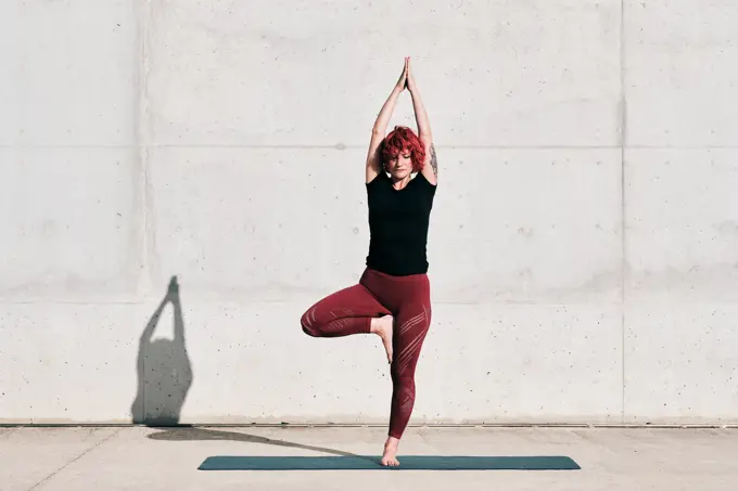 Confident barefooted female athlete in sportswear meditating while standing in vrikshasana position with arms raised and namaste on sports mat training alone on street against concrete wall in sunlight