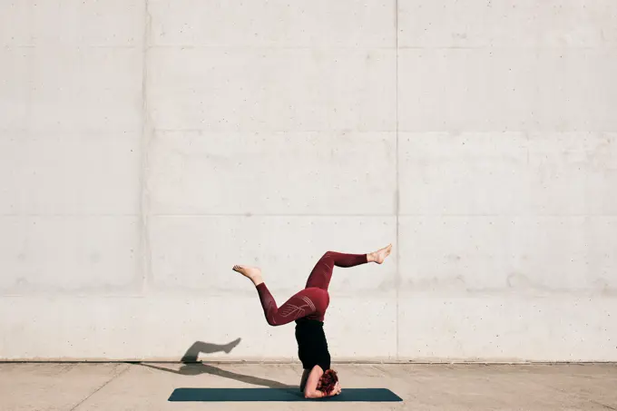 Side view of unrecognizable barefooted female athlete in activewear standing upside down in sirsasana position with legs raised in split on sports mat training alone on street against concrete wall in daytime