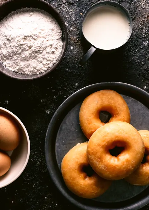 Top view of fresh doughnuts placed on rough table near various pastry ingredients and utensils in kitchen