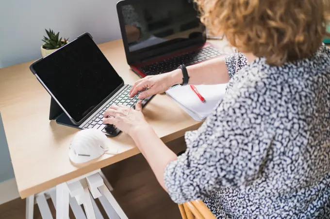 From above view of anonymous female freelancer in casual clothes typing on keyboard while working with laptops at wooden table at home