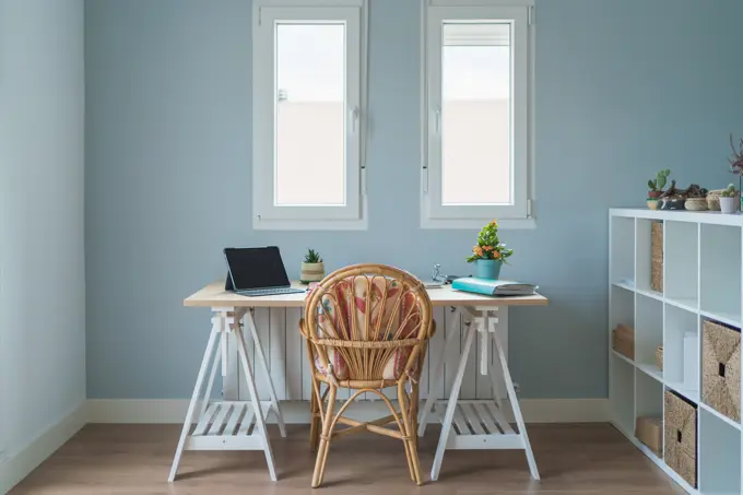 Interior of modern room and workplace with wooden desk with laptop and folder and whicker chair near wooden shelves with boxes and potted plants