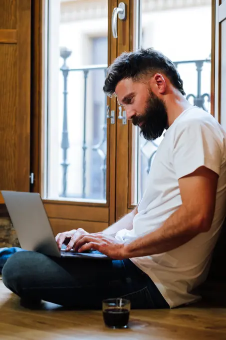 Side view of bearded man sitting on floor near mug of hot beverage and typing on laptop while working on remote project at home