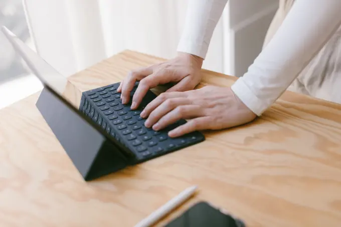 Cropped hands of unrecognizable person at wooden desk using tablet with keypad working in calm cozy office