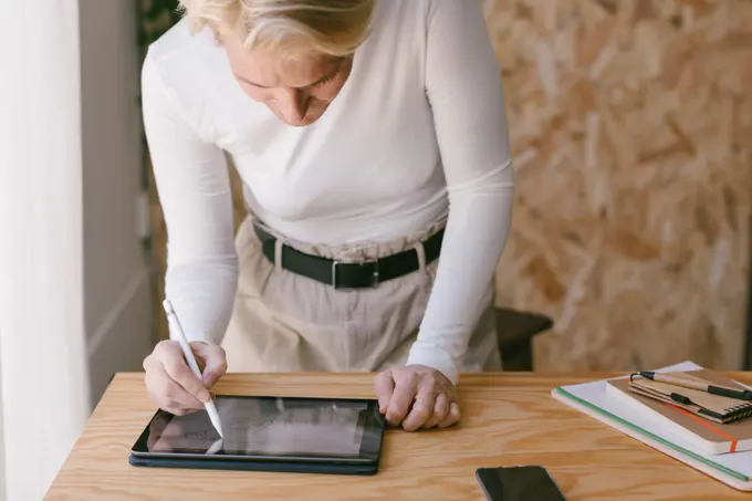 Stylish blond businesswoman bending over table and working on tablet with stylus in light wooden office