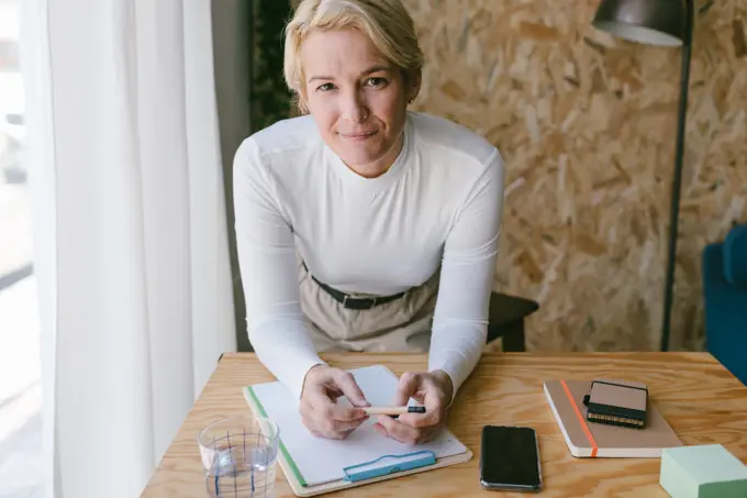 Focused adult businesswoman looking at camera while taking notes of plan on clipboard bending on wooden table in light office