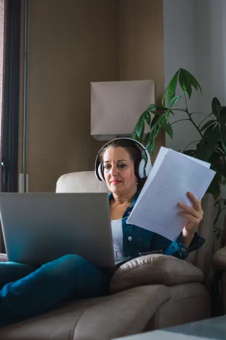 Mature woman listening to music in headphones and reading paper while sitting in armchair and doing remote job on laptop near window at home