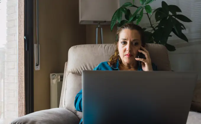 Adult woman with laptop having smartphone conversation during work at home