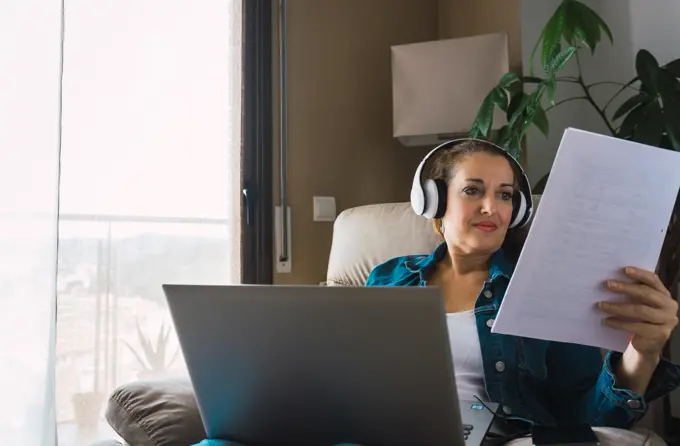 Mature woman listening to music in headphones and reading paper while sitting in armchair and doing remote job on laptop near window at home