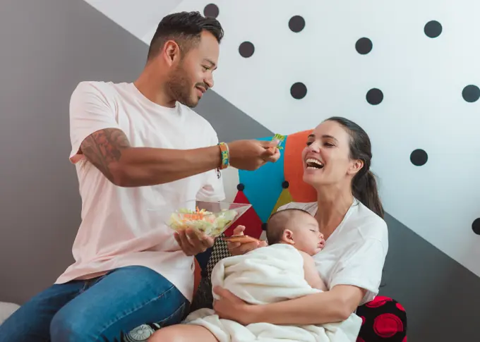 Mother with baby talking on mobile phone while sitting on armchair with casual father eating salad