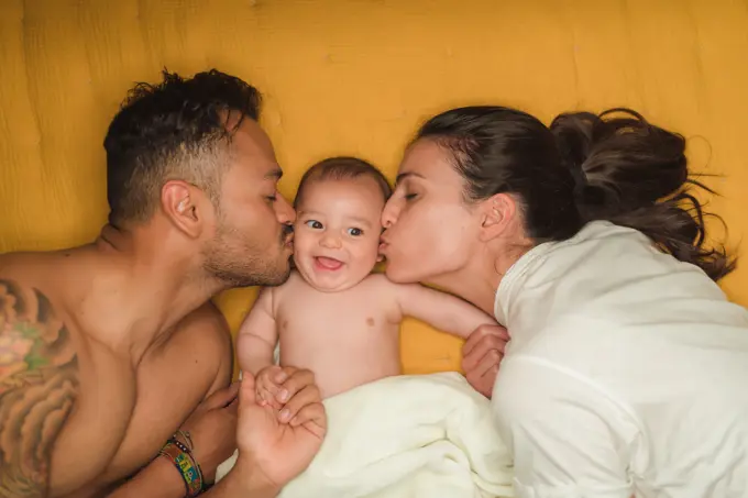 Top view of young happy parents kissing in cheeks little satisfied baby while lying on bed