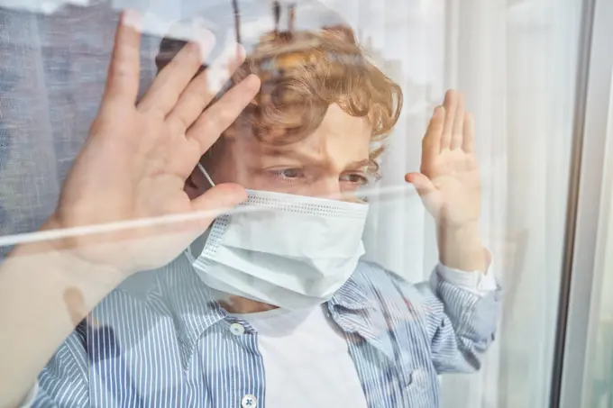 Boy in medical mask keeping hand on glass and looking through window while staying home during quarantine