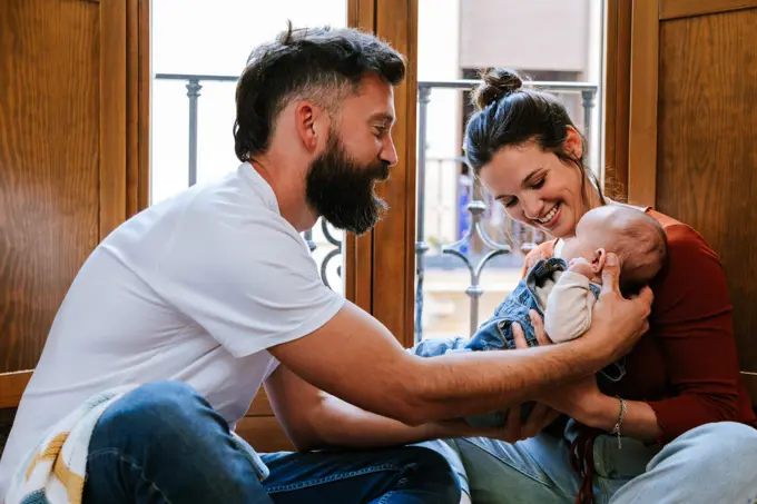 Bearded father and happy mother communicating with cute baby while sitting on floor near window at home