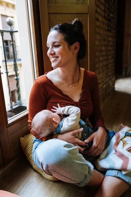 Happy mother breastfeeding baby near window