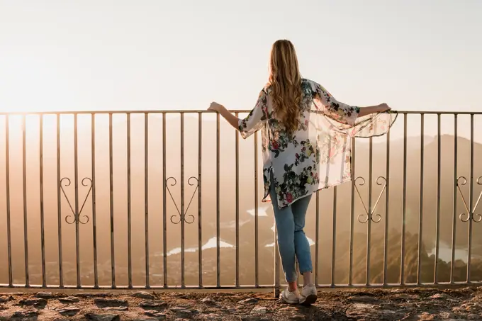 Back view of female tourist in casual clothes standing near metal fence and admiring sundown view during walk on summer day