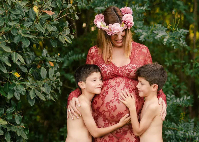 Happy mother in beautiful red dress and floral wreath smiling and embracing twin sons cuddling belly while standing near green trees in garden in summer day