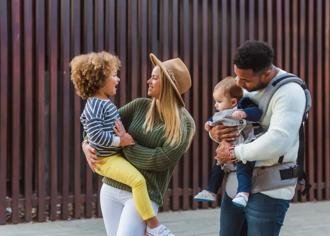 Cheerful stylish young multiracial couple with infant son and little daughter in casual wear walking together along wooden fence on city street