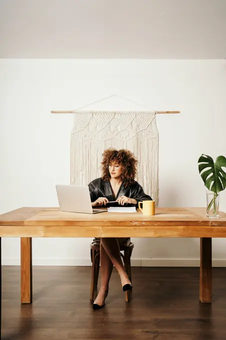 Adult lady in leather jacket and with curly hair sitting at table against macrame decoration and reading data on laptop during work on project in workplace